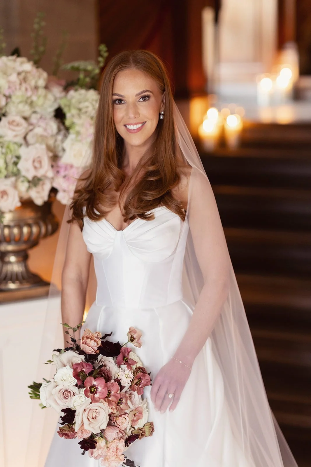 A woman in a white wedding dress holding a bouquet of soft pink and natural white flowers, standing indoors with floral arrangements and lit candles in the background.