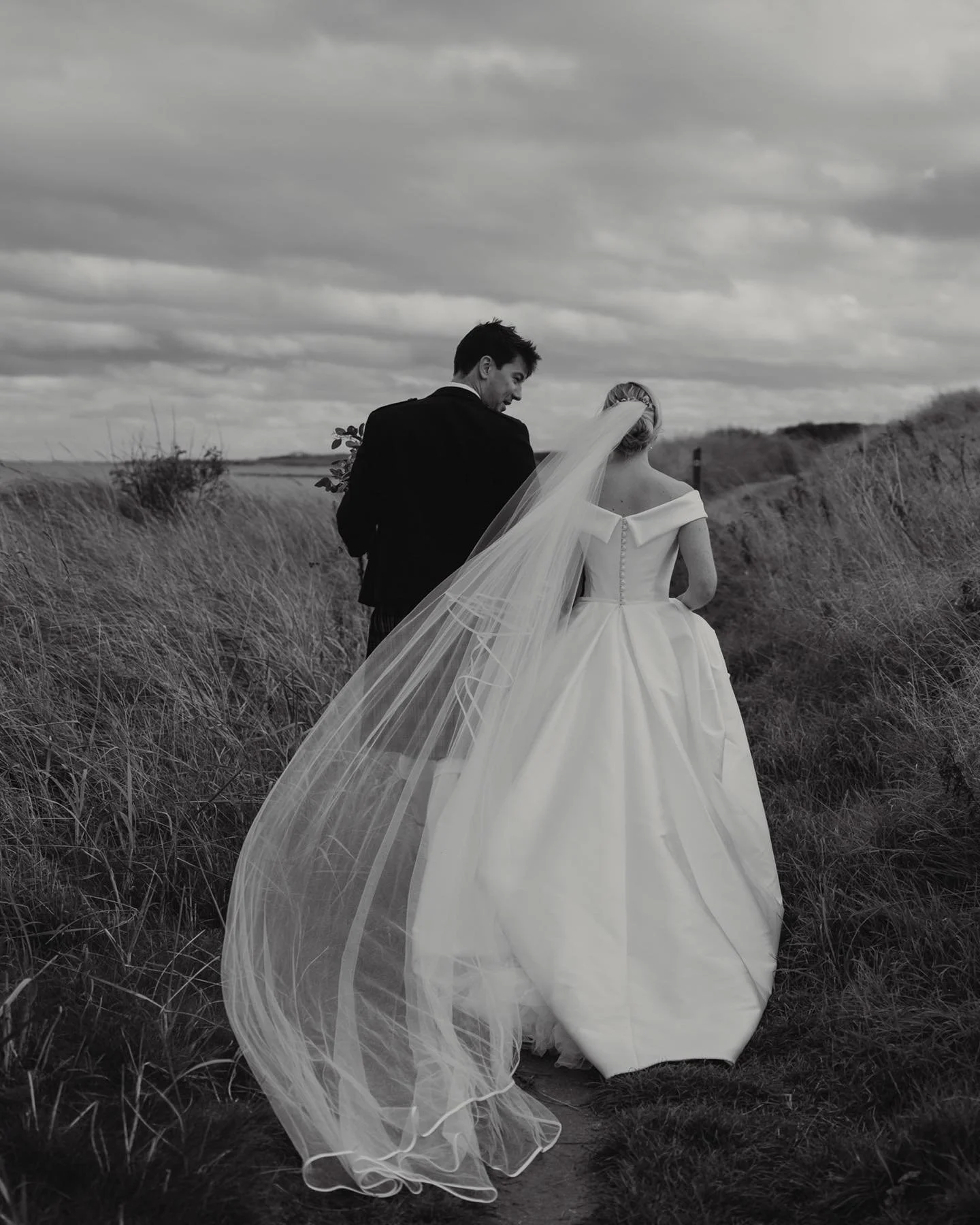 Black-and-white photo of a bride and groom walking through a grassy outdoor area, with the bride in a wedding dress and long veil, and the groom in a dark suit, under a cloudy sky.