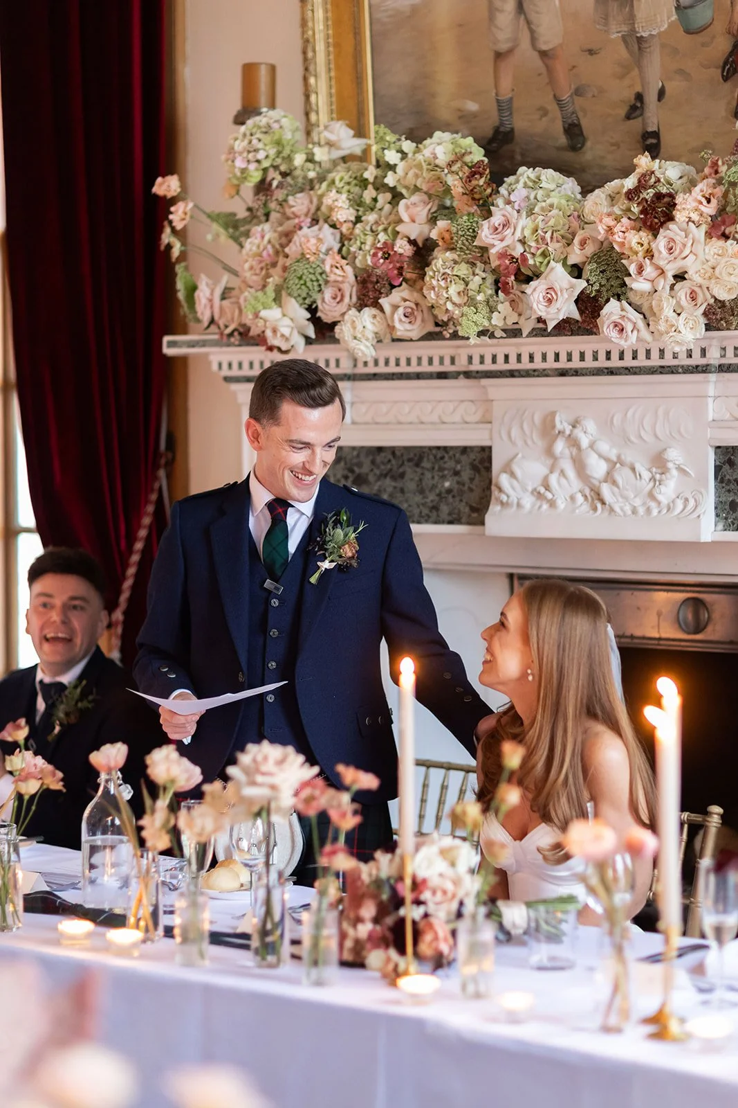 Groom in a kilt is smiling and speaking to a bride with long hair, who is looking up at him and smiling, during a wedding reception. Another man is sitting nearby, also dressed formally, with floral decorations and candles on the table, and large flo