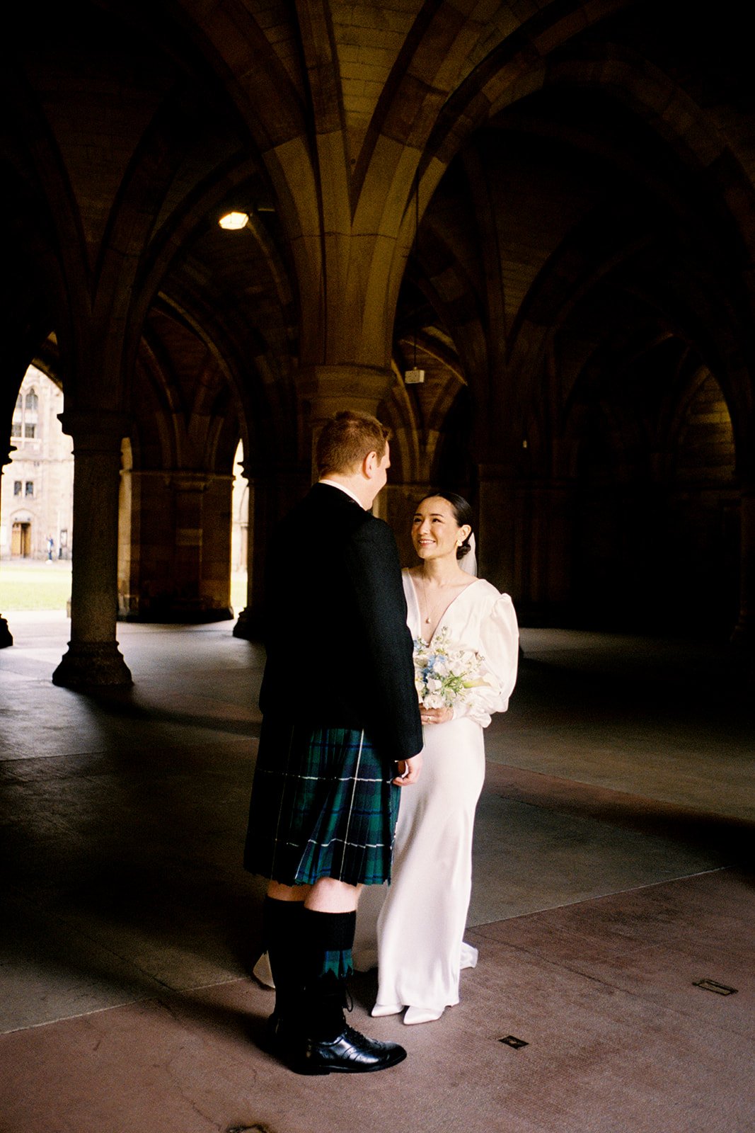 A couple dressed in wedding attire standing under a stone arched structure, facing each other and smiling, with the woman holding a bouquet of flowers.