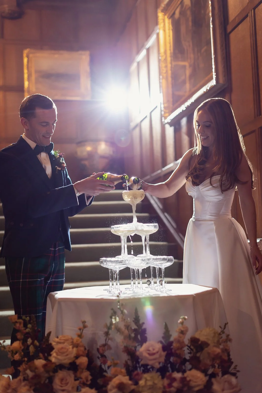A bride and groom pouring champagne into a pyramid of champagne glasses during their wedding reception with a floral arrangement at the base of the table.