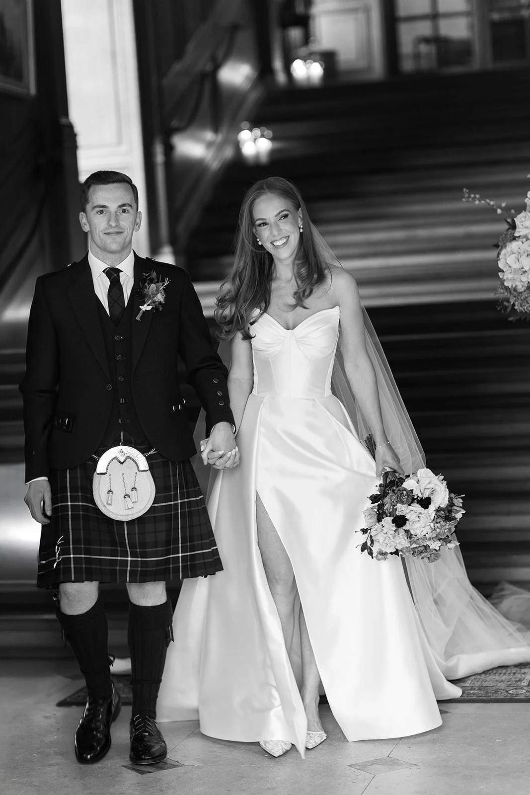 A black-and-white photo of a newlywed couple holding hands and walking down a staircase. The groom is dressed in a kilt and the bride is wearing a strapless wedding gown with a thigh-high slit and a veil, holding a bouquet of flowers.