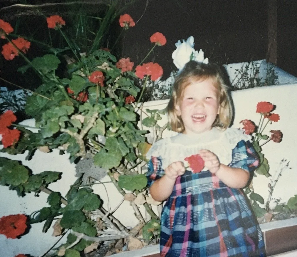 A young girl smiling and holding a red flower, standing next to a large plant with red flowers.
