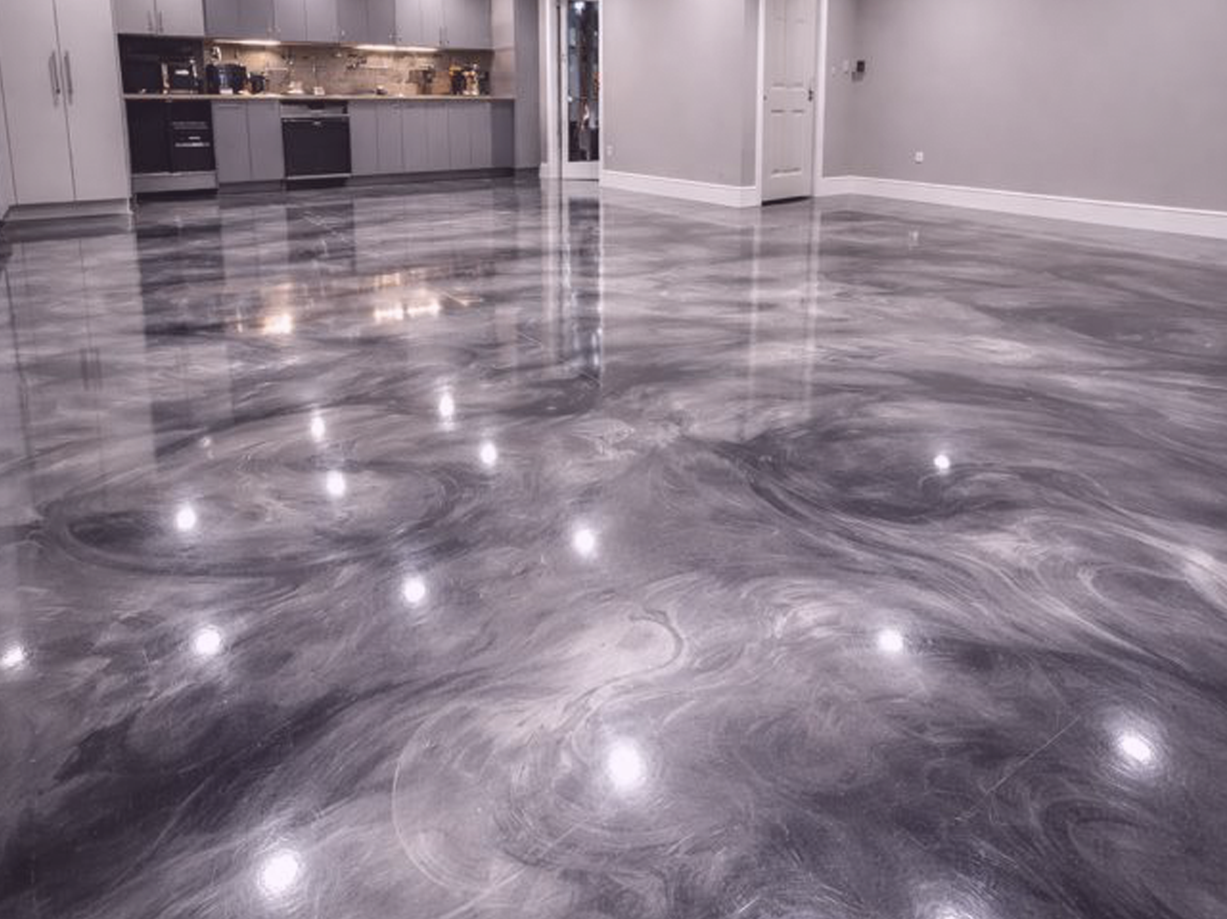 Polished metallic epoxy floor in an open kitchen and living area with gray cabinets, a white door, and white baseboards.