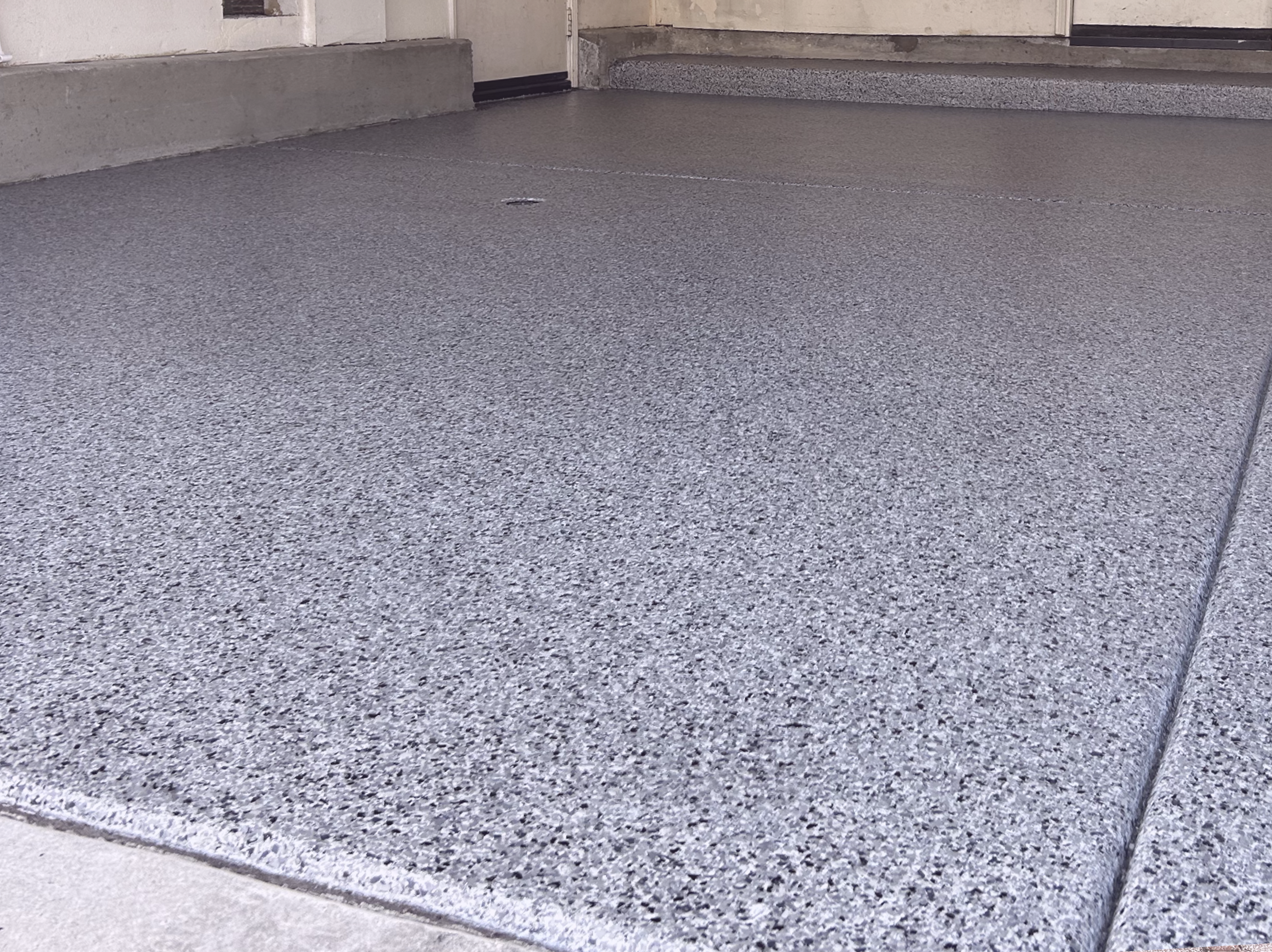 A textured gray concrete epoxy full color chip system porch or sidewalk with a drain cover near the center, adjacent to a building with beige siding and steps.