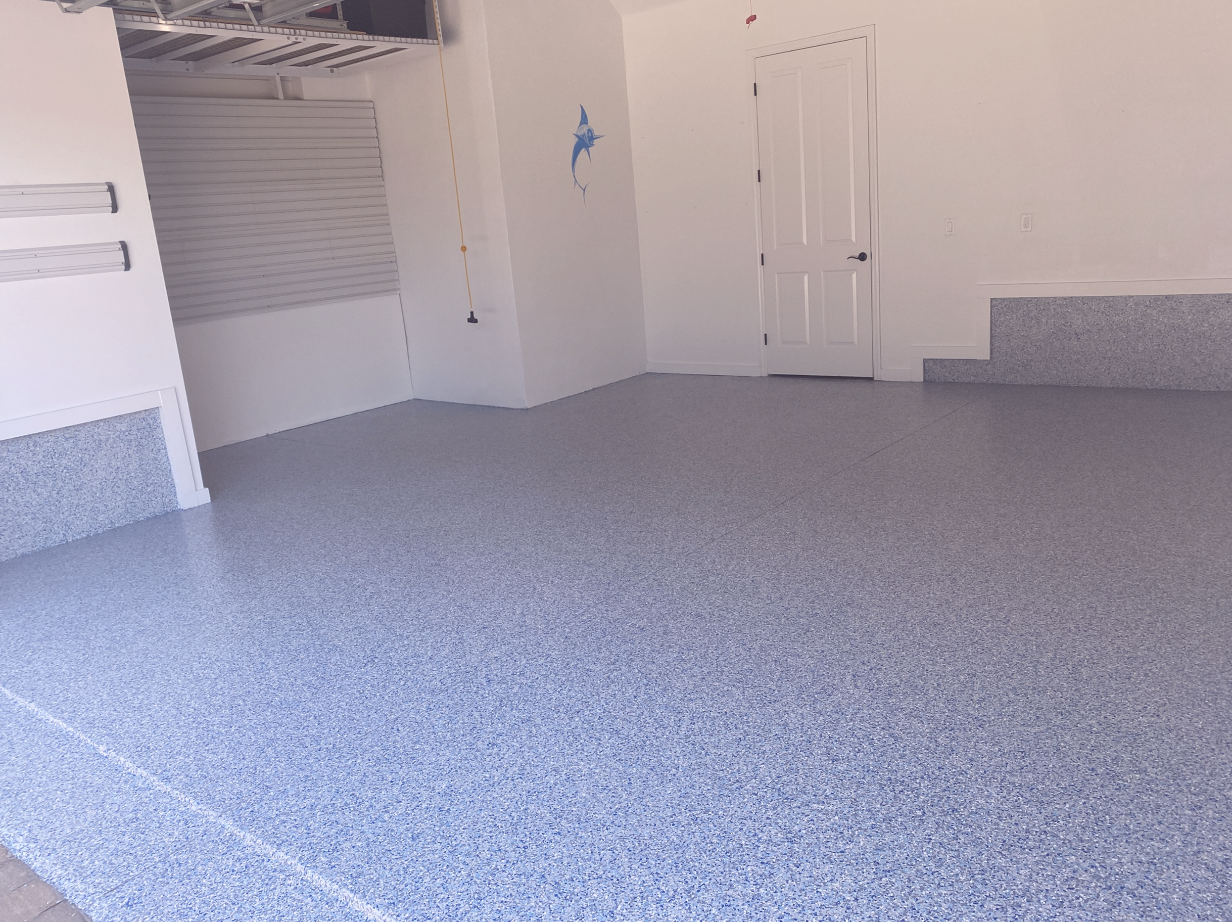 Empty garage with white walls, gray speckled epoxy flooring, a closed white door, wall-mounted storage and hooks, and a marlin decal on the wall.