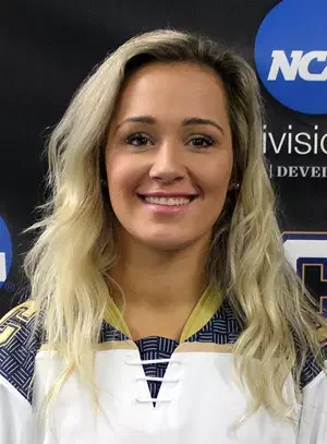 Young woman with long blonde hair wearing a sports jersey, smiling, standing in front of a black background with a partially visible NCAA logo.