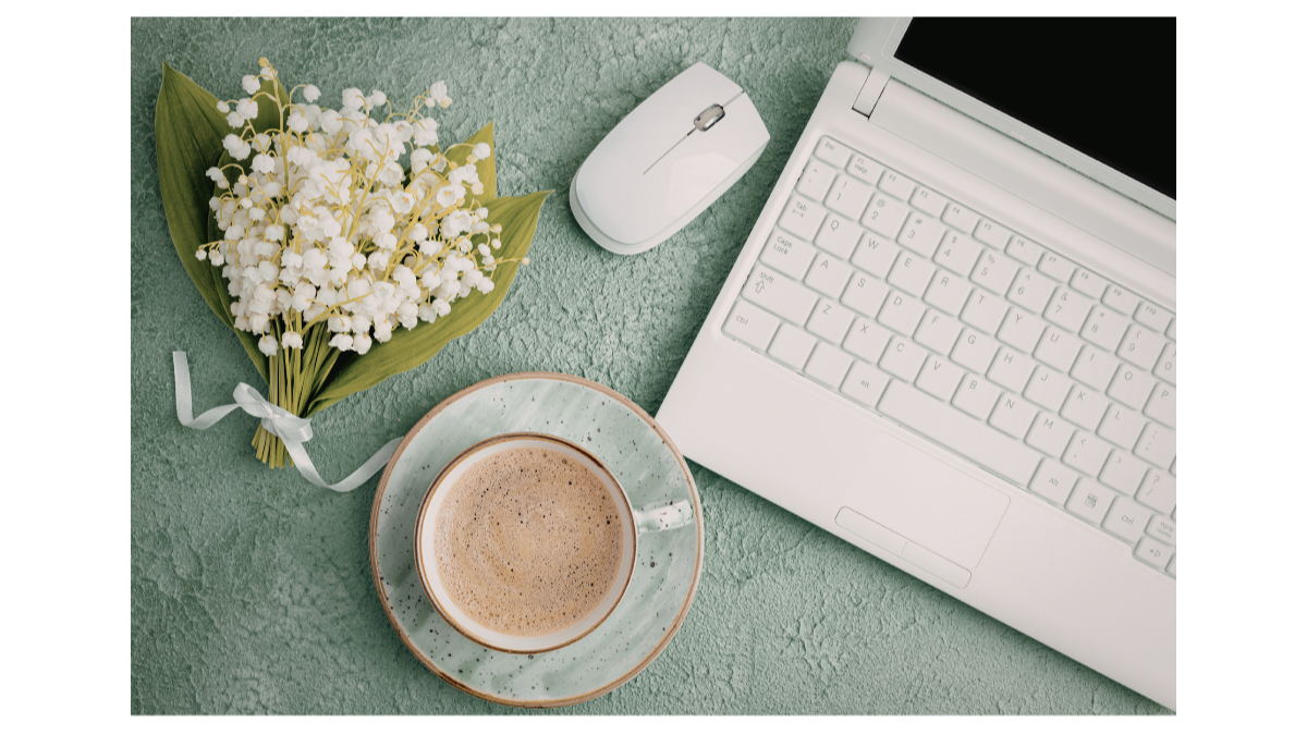 A top view of a workspace with a bouquet of white flowers, a white computer mouse, a cup of coffee on a saucer, a white laptop, all on a textured green surface.