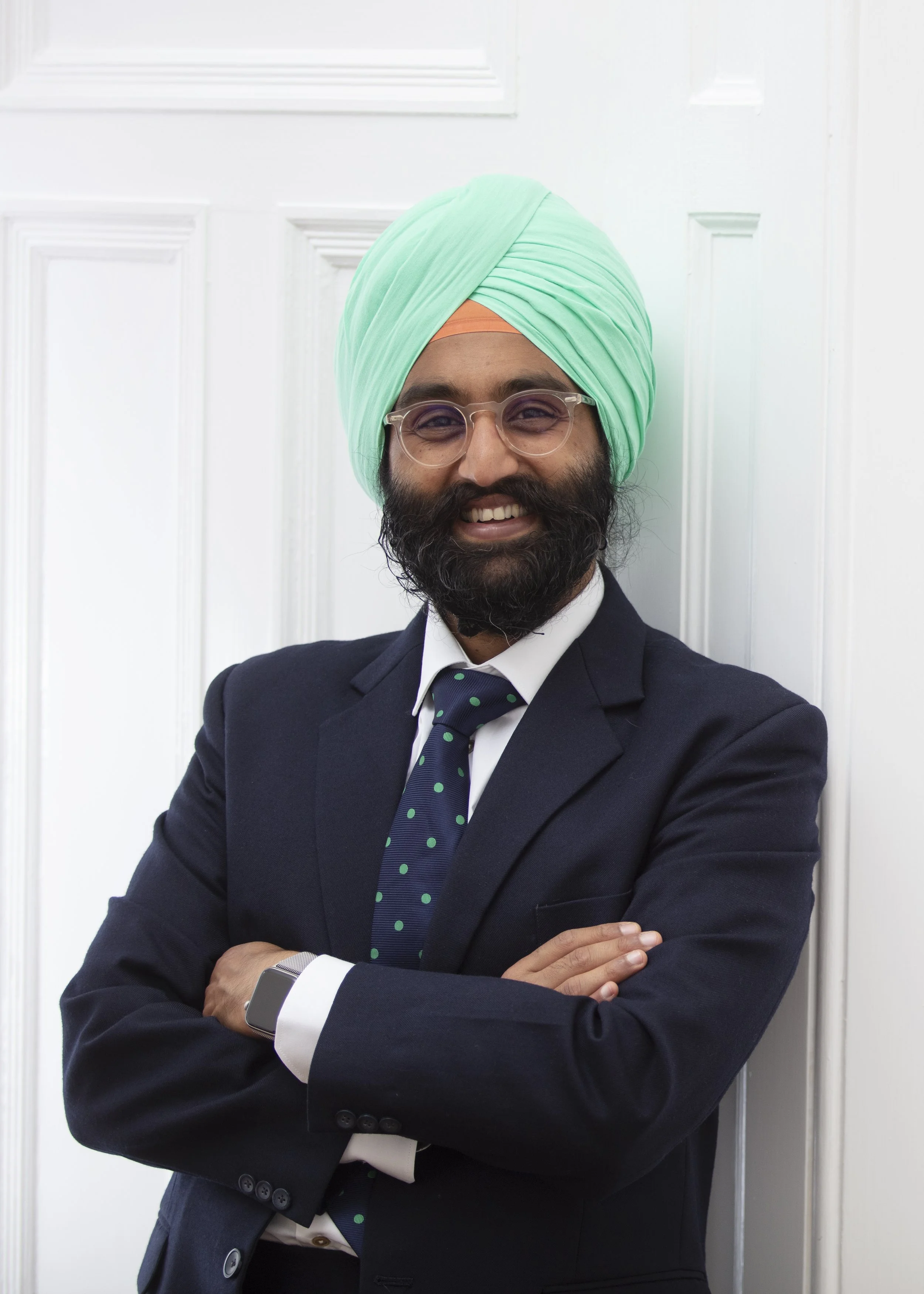 A man with a beard dressed in a business suit and tie, wearing a mint green turban, standing in front of a white paneled wall, smiling with arms crossed.