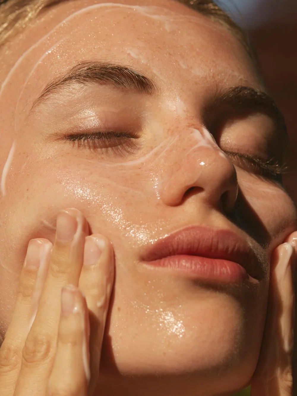 Close-up of a woman with her eyes closed, applying a facial coating or mask, with a serene expression.