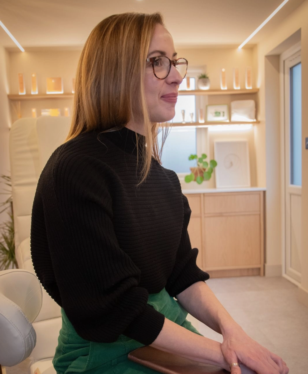 A woman with glasses and long red hair sitting in a modern, well-lit room with light-colored walls, shelves with decorative items, and a window.