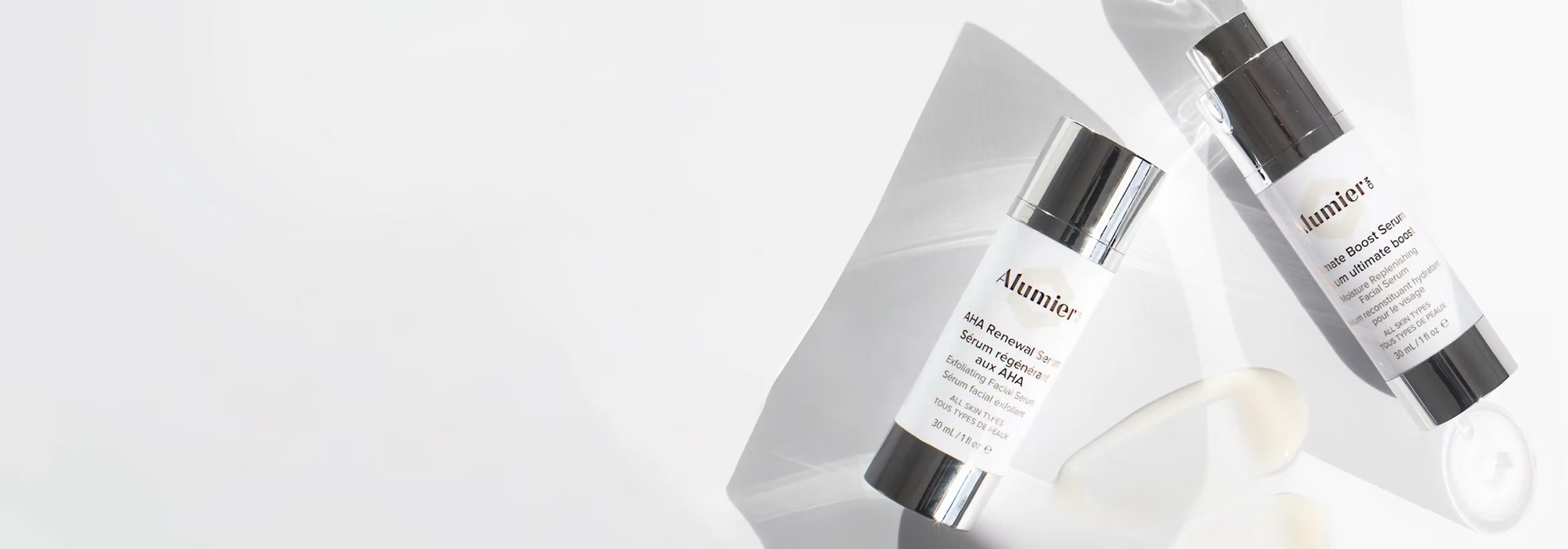 Two white Alumier skincare serum bottles with metallic caps labeled 'Alumier' and 'AHA Renewal Serum' and 'Skin Brightening Serum' placed on a diagonal shadowed surface.