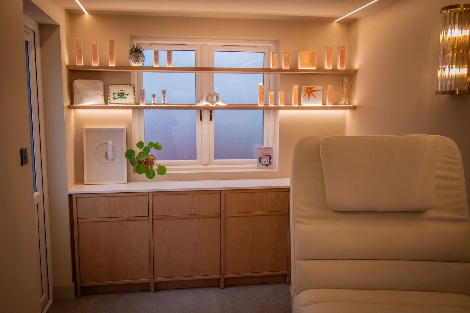 Finer Medical Aesthetics studio with a window, wooden shelves with decorative items, a potted plant, and a beige sofa.