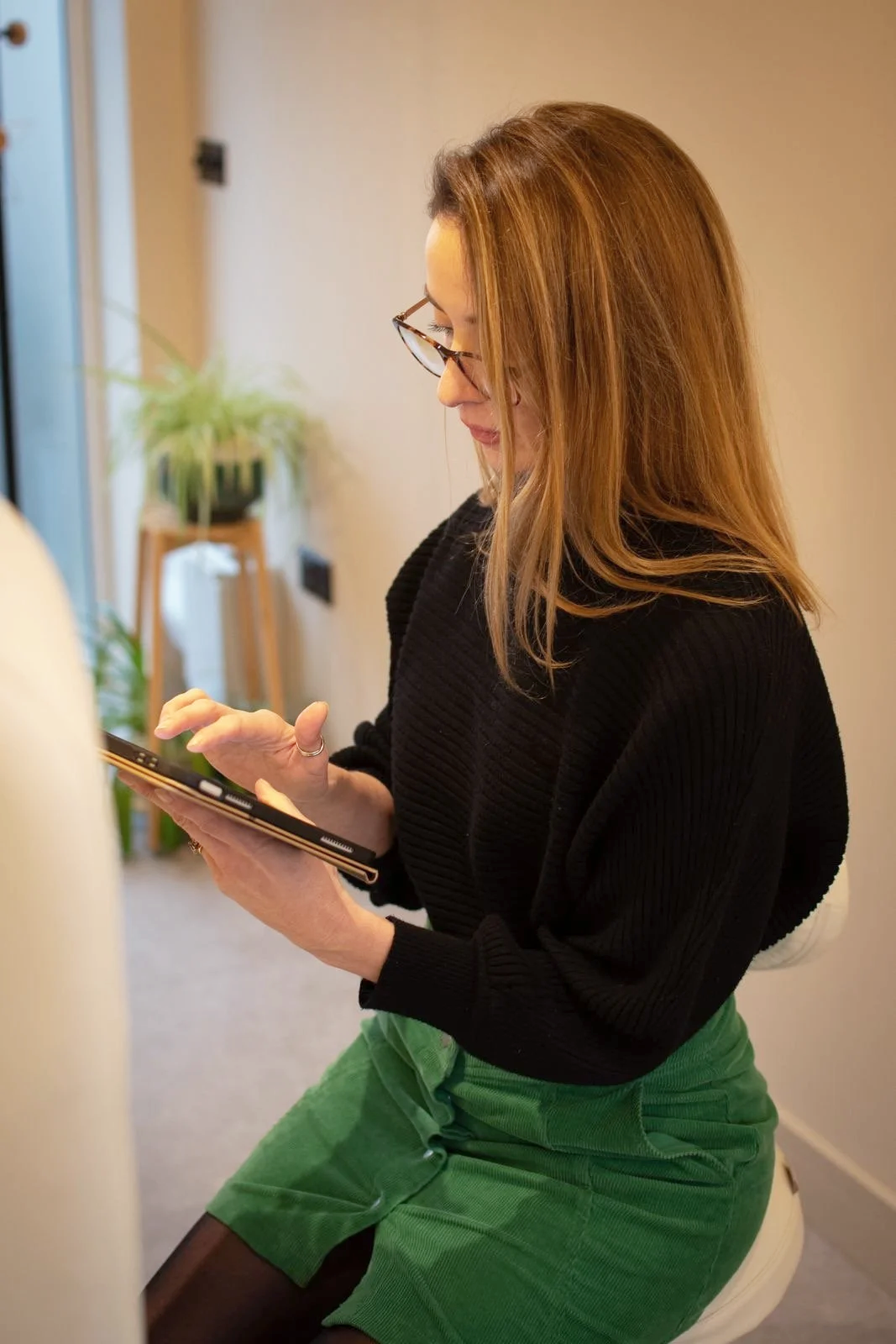 A woman with blonde hair and glasses sitting on a stool in a room, looking at and using her smartphone, with potted plants and a door in the background.