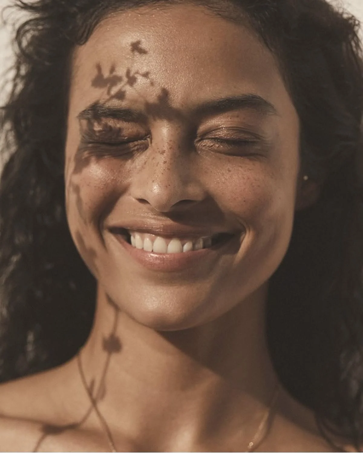 Close-up of a woman's smiling face with perfect skin, closed eyes, curly hair, and cast shadows on her forehead.