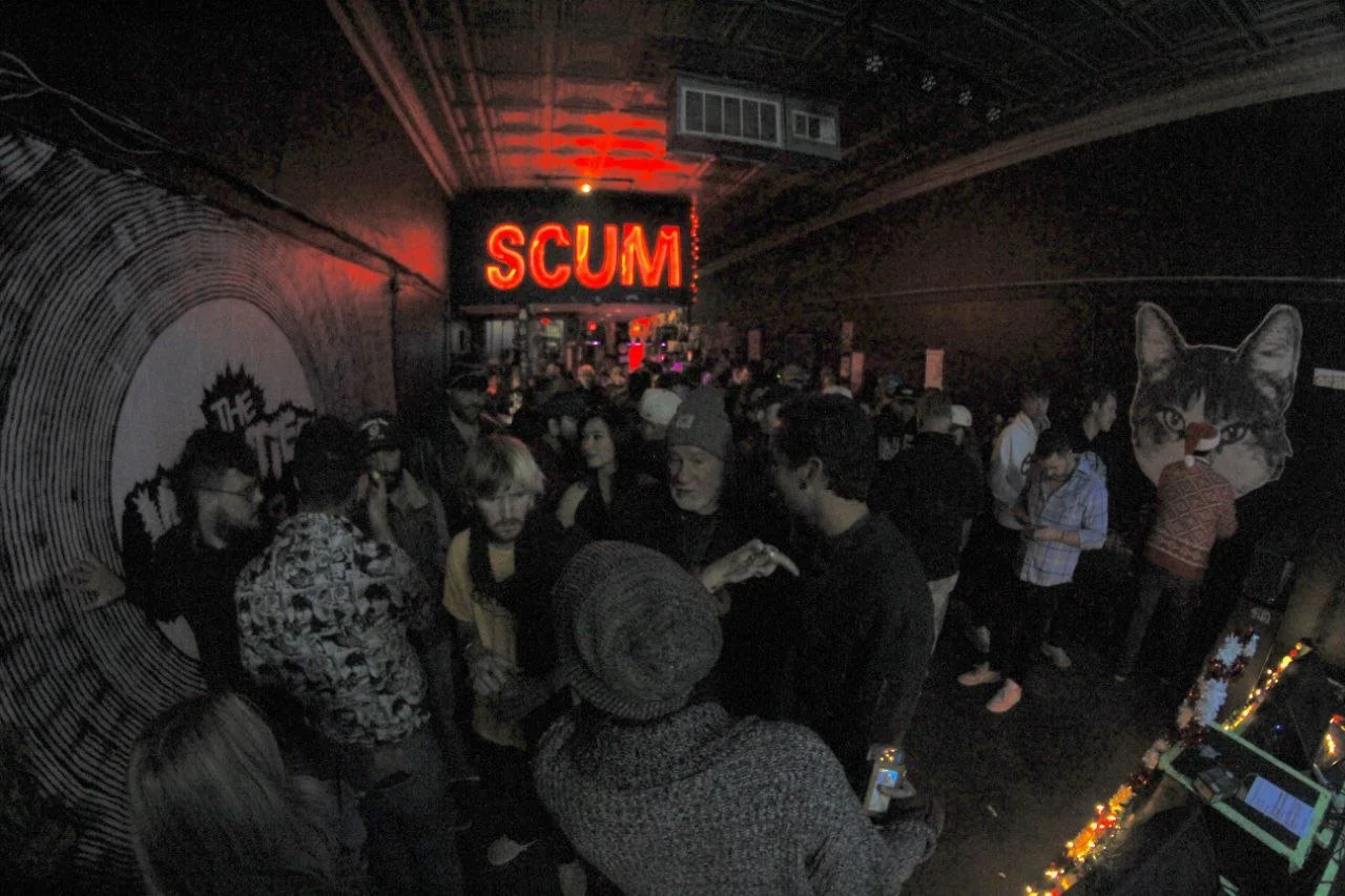 People gathered in a dark nightclub with a neon sign that reads 'SCUM' on the wall.