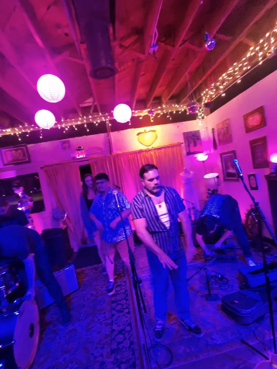 A band performing on a colorful, neon-lit stage with purple and pink lighting, paper lanterns, and string lights on the ceiling. Several musicians are standing near microphones, with a drum set visible on the left.