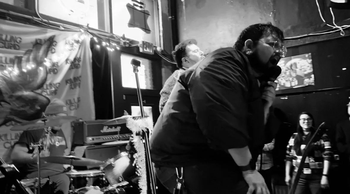 In a black and white image, a vocalist passionately sings into a microphone in a music venue, accompanied by a drummer and a keyboardist, with audience members watching, and stage decor including balloons and string lights.