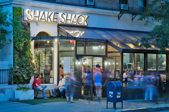 Outdoor patio at Shake Shack with people sitting at tables, dining and socializing, near a building with greenery and city buildings in the background by Davella Studios.