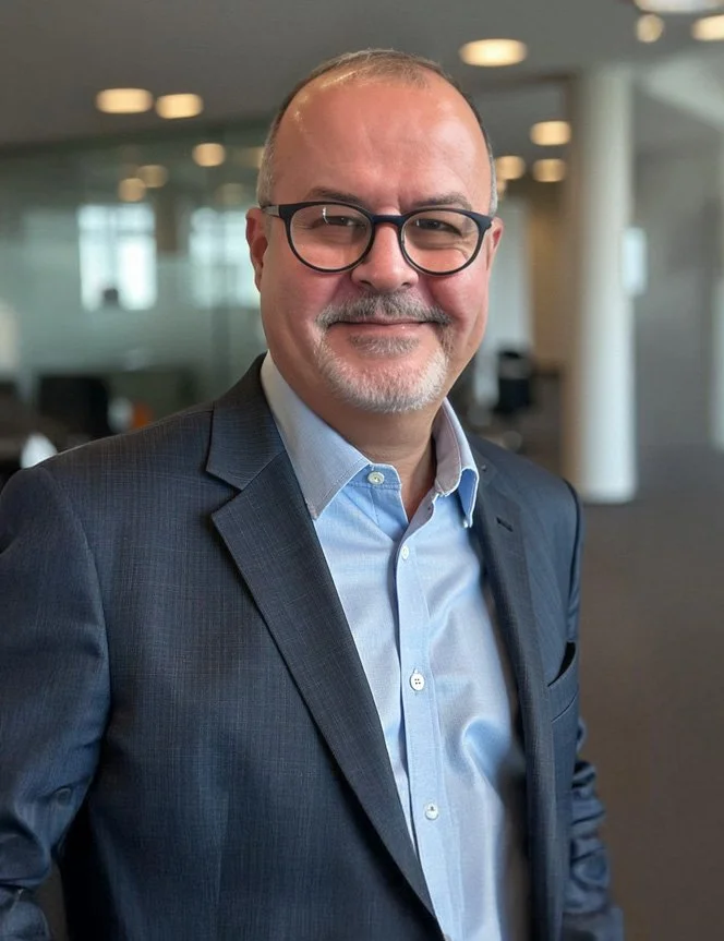 Mauricio Porto wearing glasses, a blue dress shirt, and a dark blazer. He is smiling in an indoor office setting with blurred background.