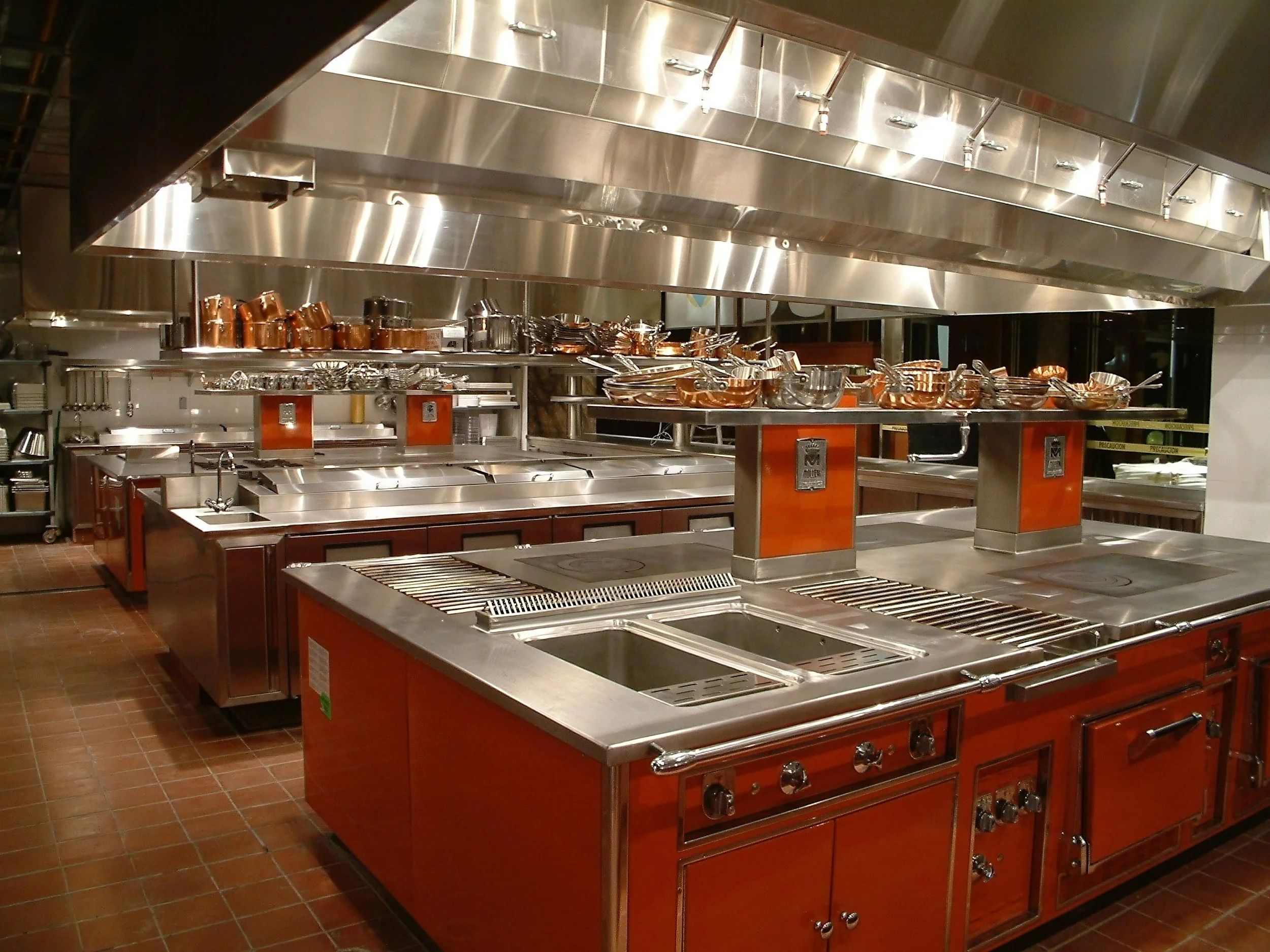 A professional stainless steel commercial kitchen with multiple cooking stations and copper pots on shelves at Le Cirque by Davella Studios.