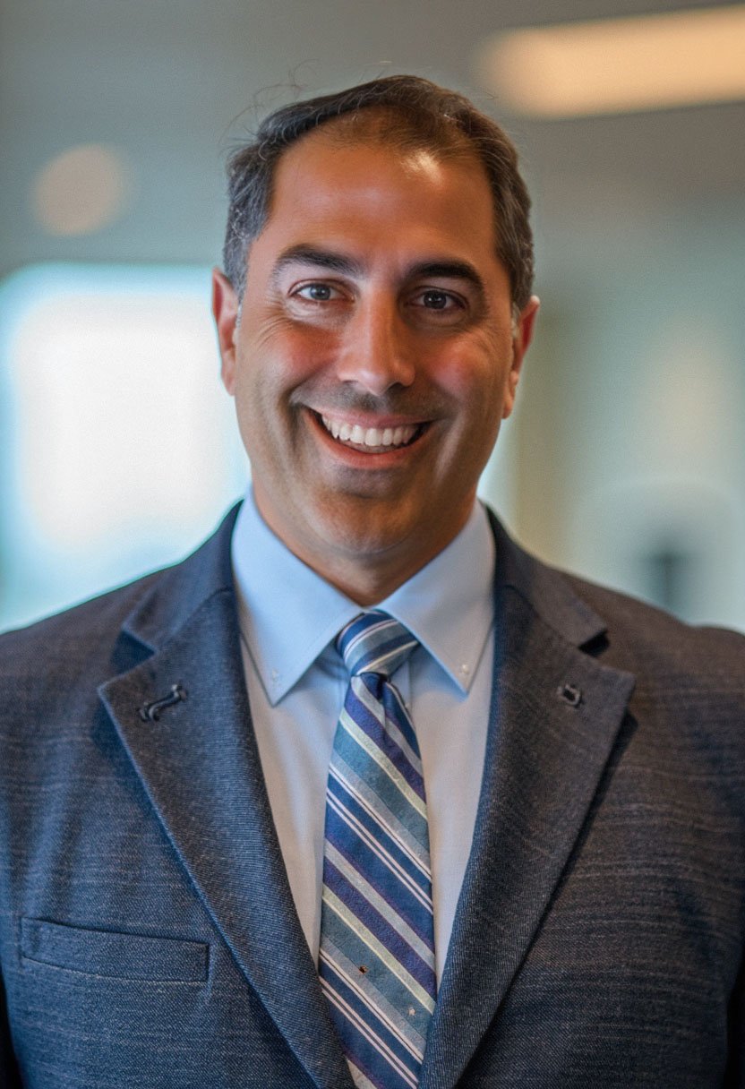 Portrait of a smiling man in a business suit with a blue-striped tie, standing indoors with blurred background.