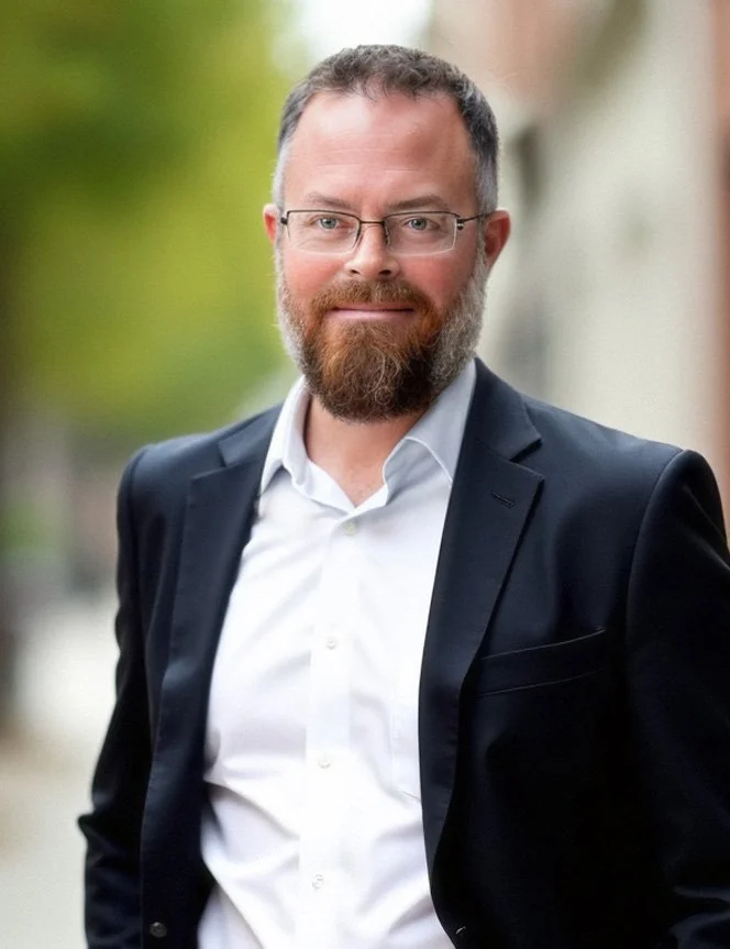 Matt Daly with glasses and a beard, dressed in a black blazer and white shirt, standing outdoors with blurred greenery in the background.