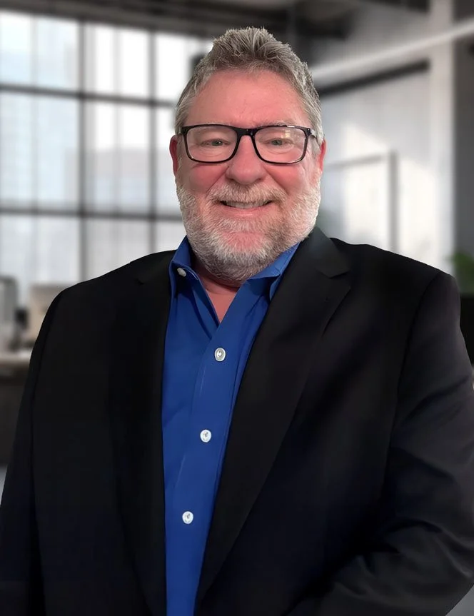 Eric McConnell with gray hair, beard, and glasses, wearing a blue shirt and black blazer, standing in a modern office with large windows.
