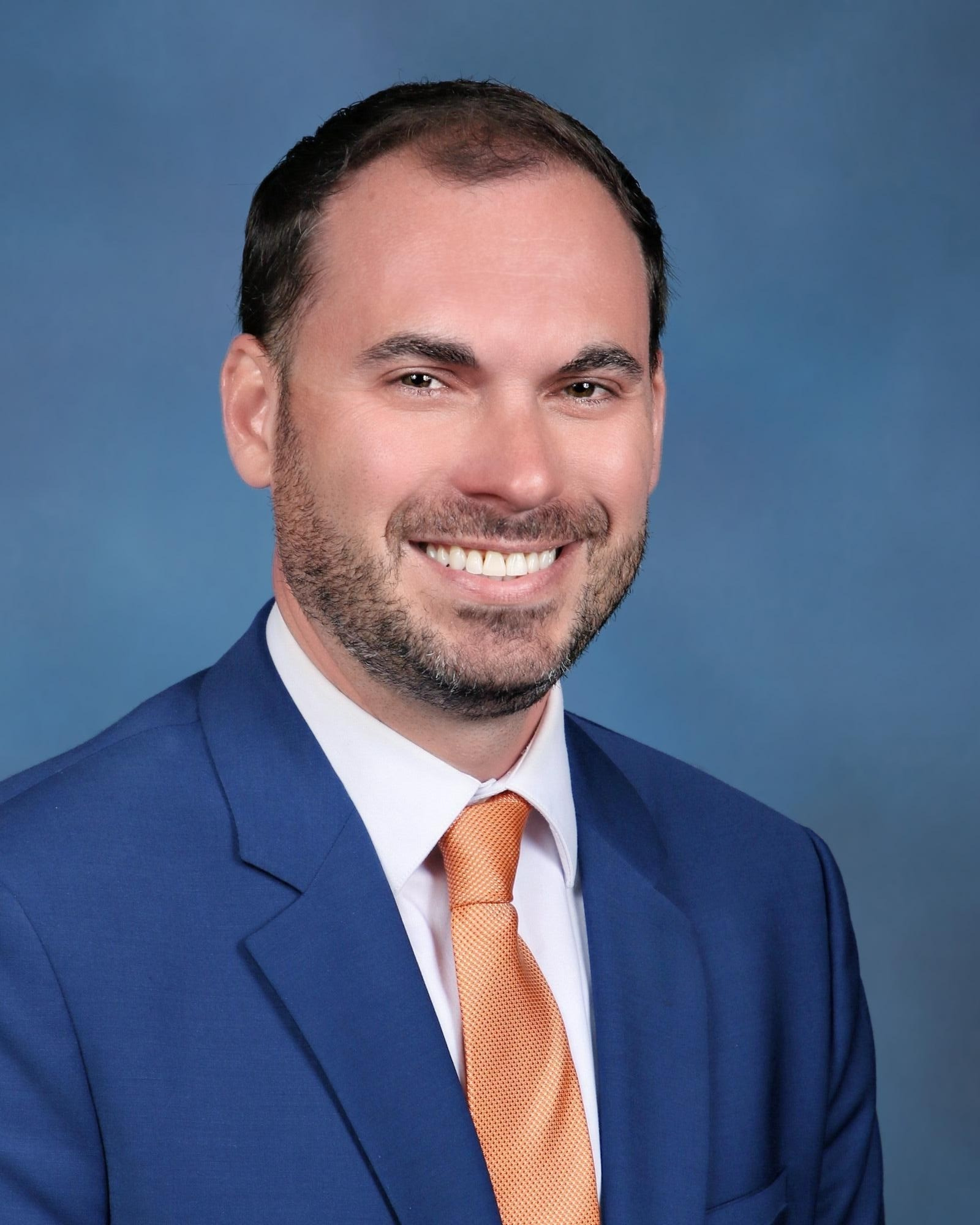 Headshot of a man in a blue suit with an orange tie, smiling, against a blue background.