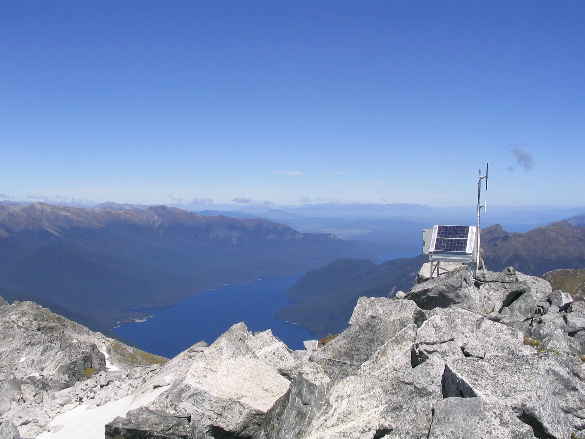 A mountain landscape with a clear blue sky, a lake in the valley, and a rocky summit with a solar panel and weather station equipment.