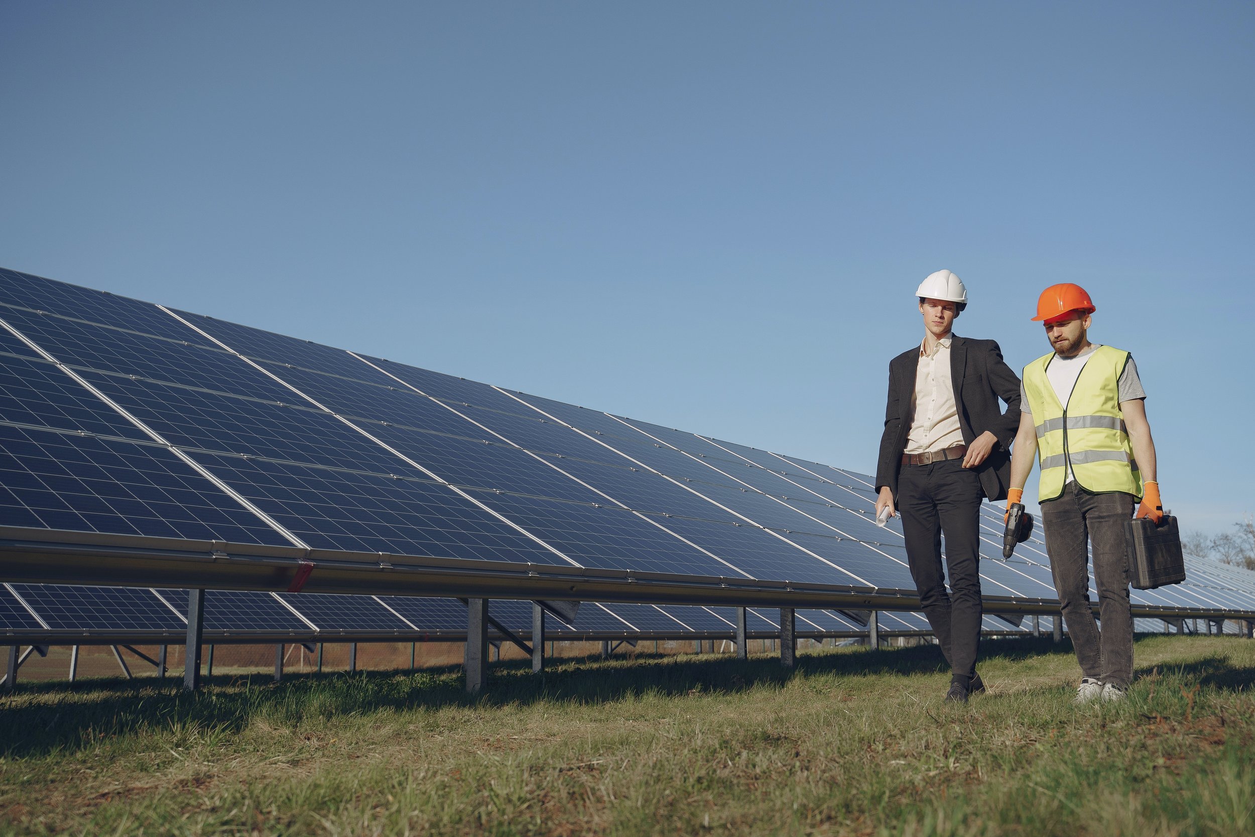 Two men in safety helmets and vests inspecting solar panels in a sunny outdoor setting.