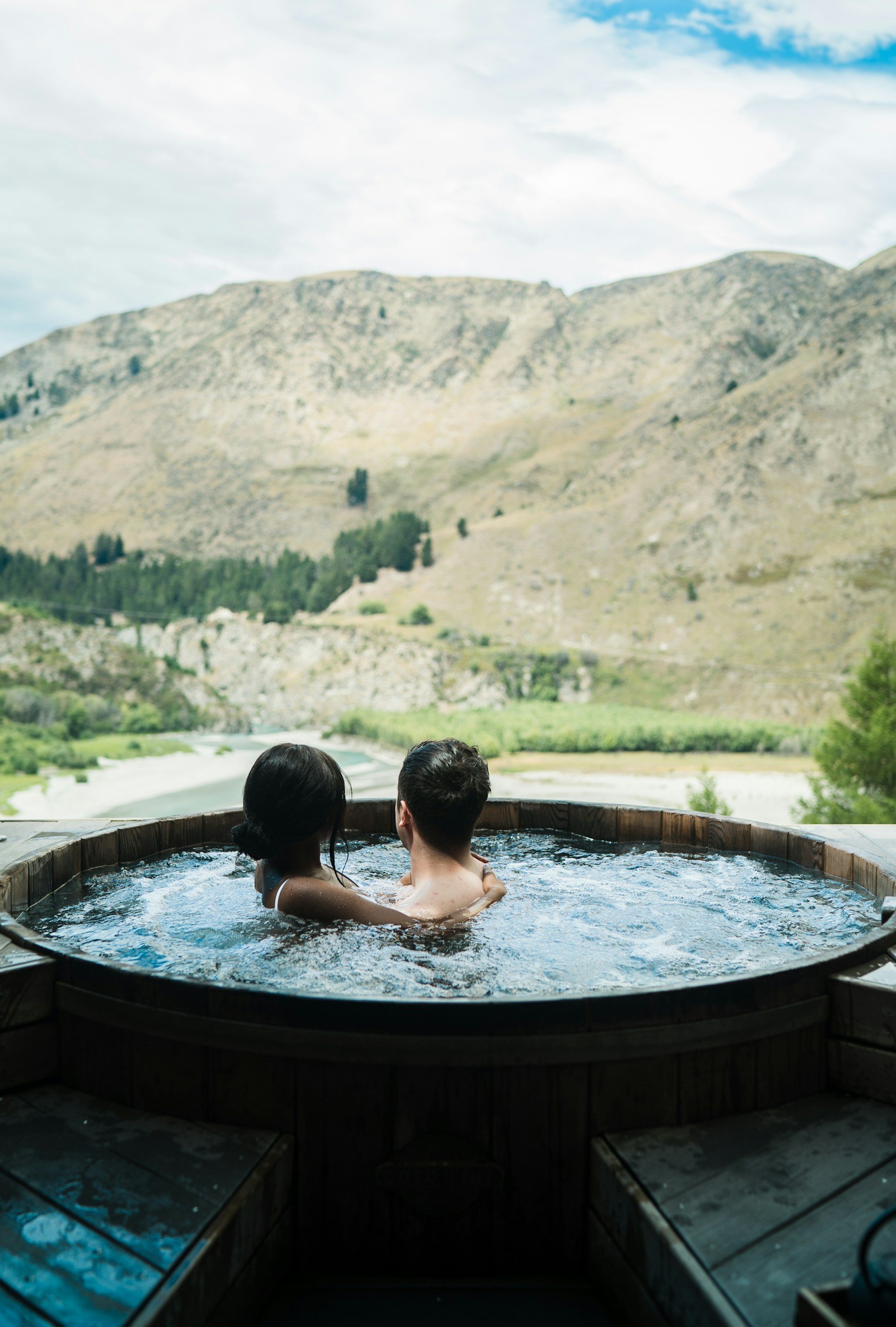 Two people sitting in a hot tub overlooking a mountainous landscape with a river.