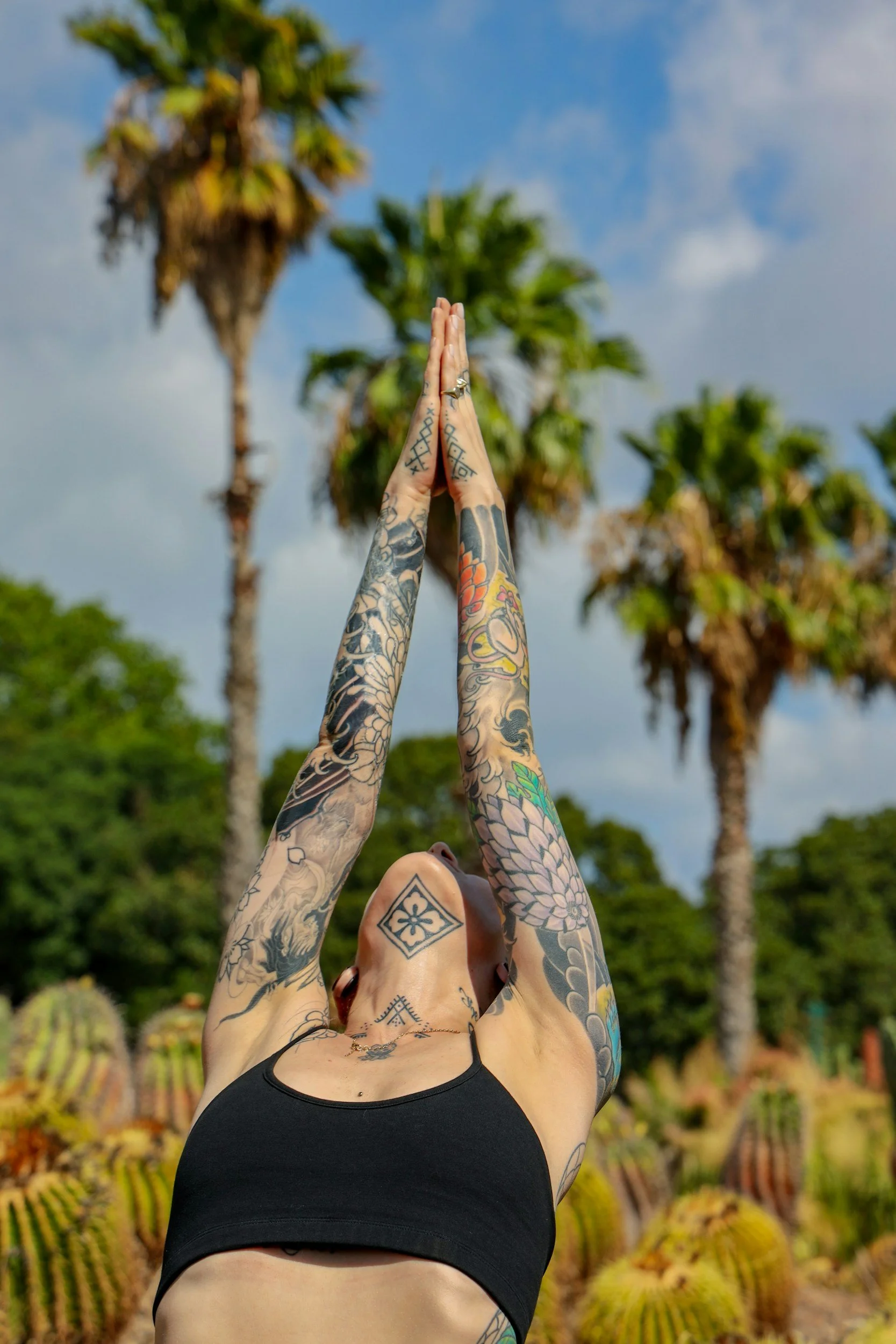 A tattooed woman in a black sports bra practicing yoga outdoors, with her hands pressed together above her head, looking upward. She is surrounded by a desert landscape with cacti and tall palm trees, under a partly cloudy sky.