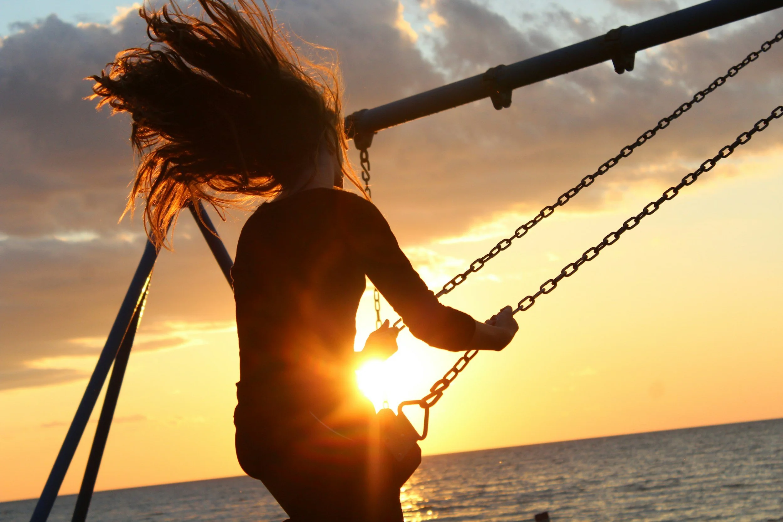 A girl swinging on a swing set at sunset along the beach, with her hair blowing in the wind and the sun partially hidden behind her, casting a warm glow over the scene.