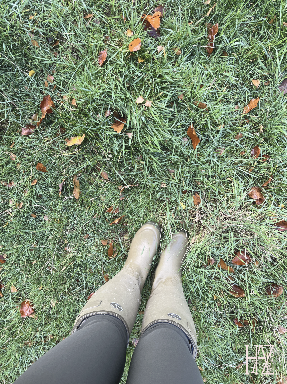 The photo shows a top-down view of a person's legs wearing black leggings and beige rain boots standing on green grass with fallen brown leaves.