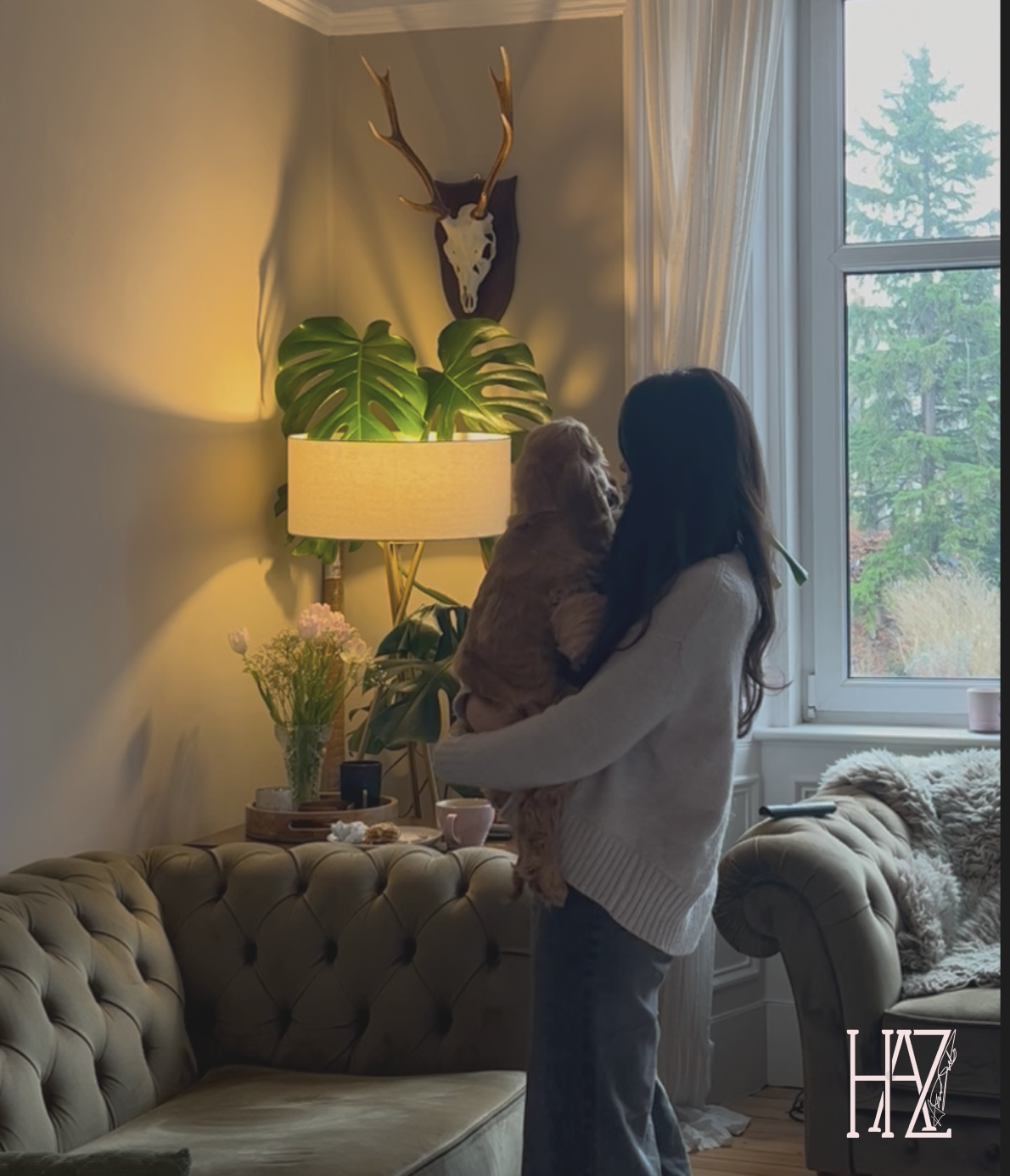 A woman holding a small dog in a cozy living room with natural light coming through a large window. The room features a beige tufted sofa, a tall lamp with a shade, a large leafy plant, and a mounted deer skull with antlers on the wall.