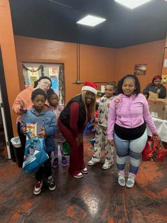 Group of children and women gathered indoors, some holding gifts, with one woman wearing a Santa hat, in a room decorated for the holiday season.