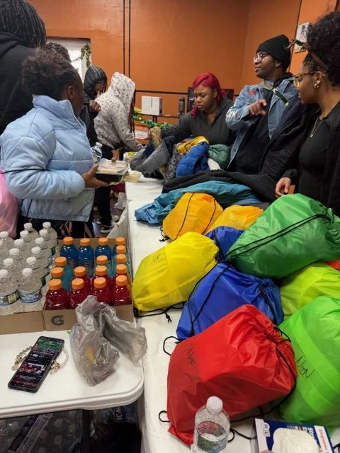 People gathering around a table with backpacks, water bottles, and snacks in a room with orange walls, likely at a community event or donation drive.