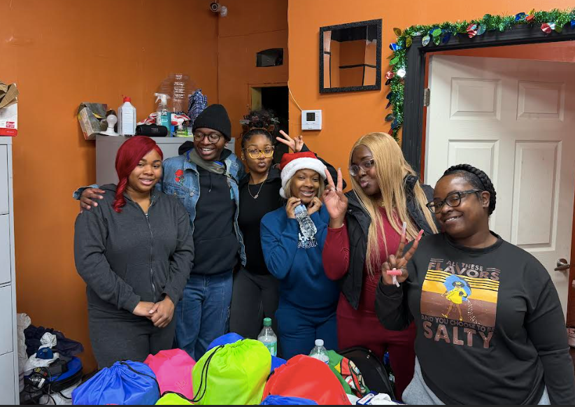 Six women standing together in a room decorated for Christmas, some making peace signs, with Christmas gear and colorful bags on the table in front of them.