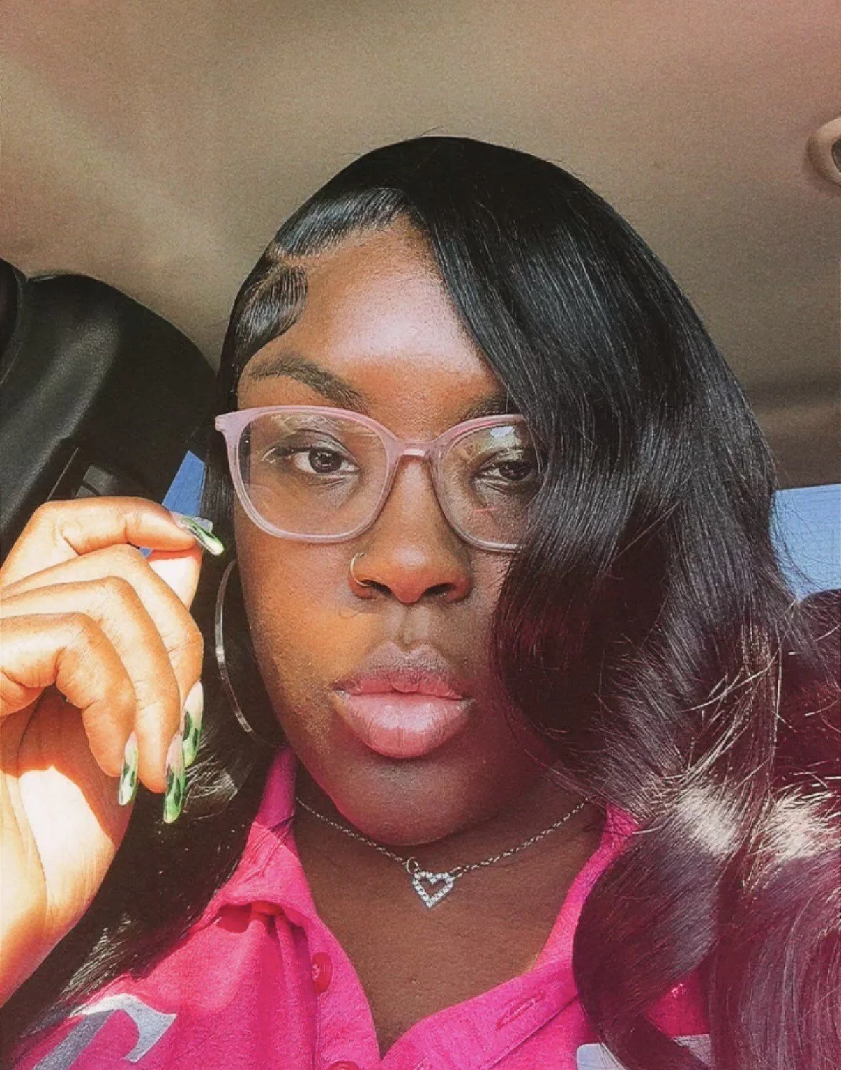 A woman with dark hair, outlined in a side part and styled in loose waves, wearing pink glasses, hoop earrings, a star-shaped necklace, and a pink shirt, holding her glasses with her left hand.