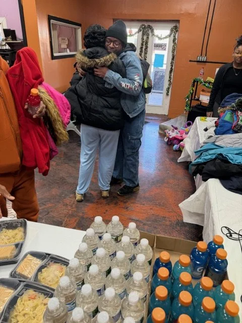 Two women hugging in a warm embrace inside a room, with a group of people around, some partially visible. There are tables with bottled drinks and food in the foreground, and a door with a window behind them.