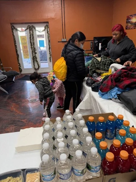 Two women shopping for clothes with three children inside a cozy store decorated for the holidays, with water bottles, energy drinks, and snacks on a table in the foreground.