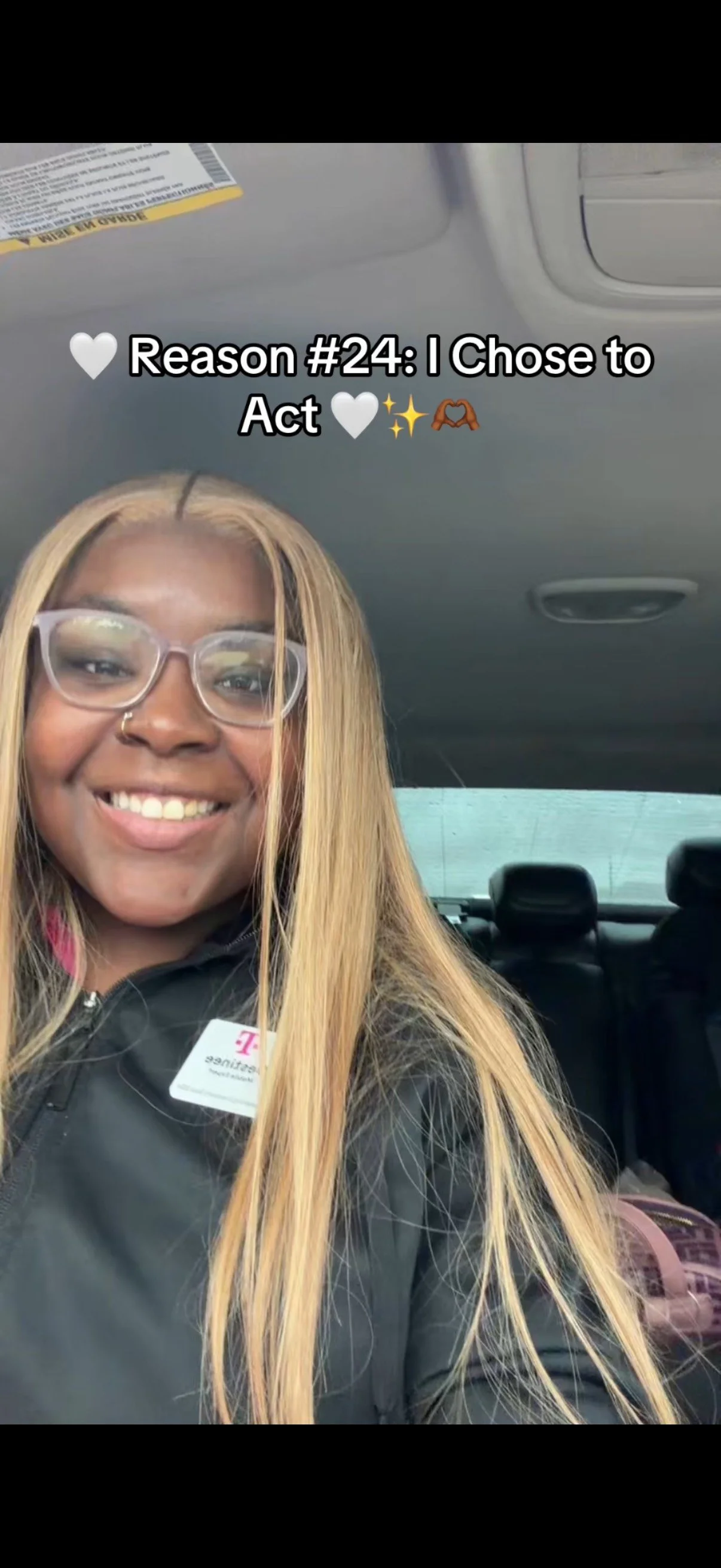 A young woman with blonde hair, glasses, and a nose ring smiling inside a car, wearing a black jacket with a name tag.