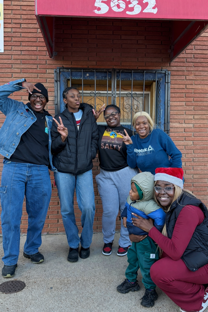 Five women and a child standing in front of a brick building with a red awning. One woman wears a Santa hat, and the child is wearing a green hat. Everyone is smiling and making peace signs.