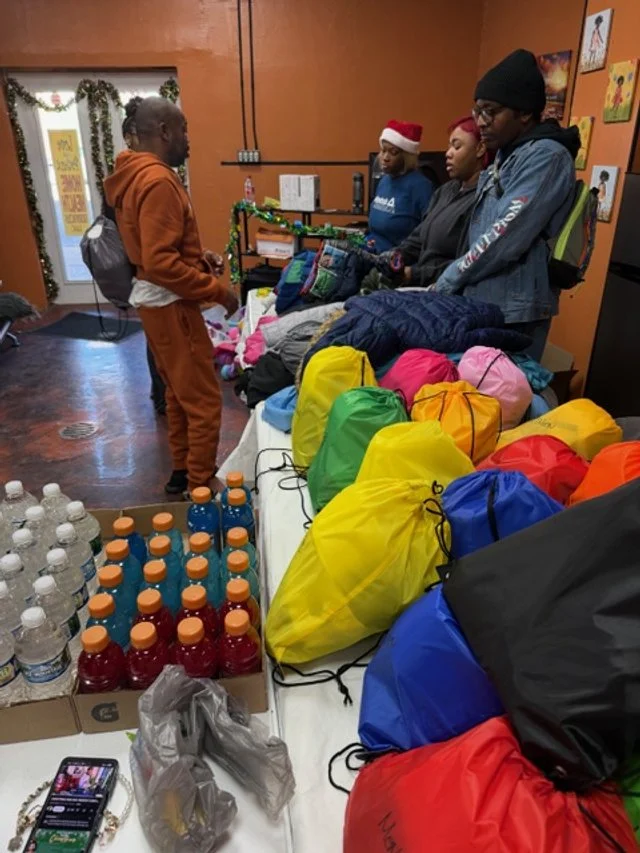People standing behind a table filled with packs of colorful gear, bottled water, and red drinks. One person is speaking to three others, two of whom are wearing holiday-themed hats.