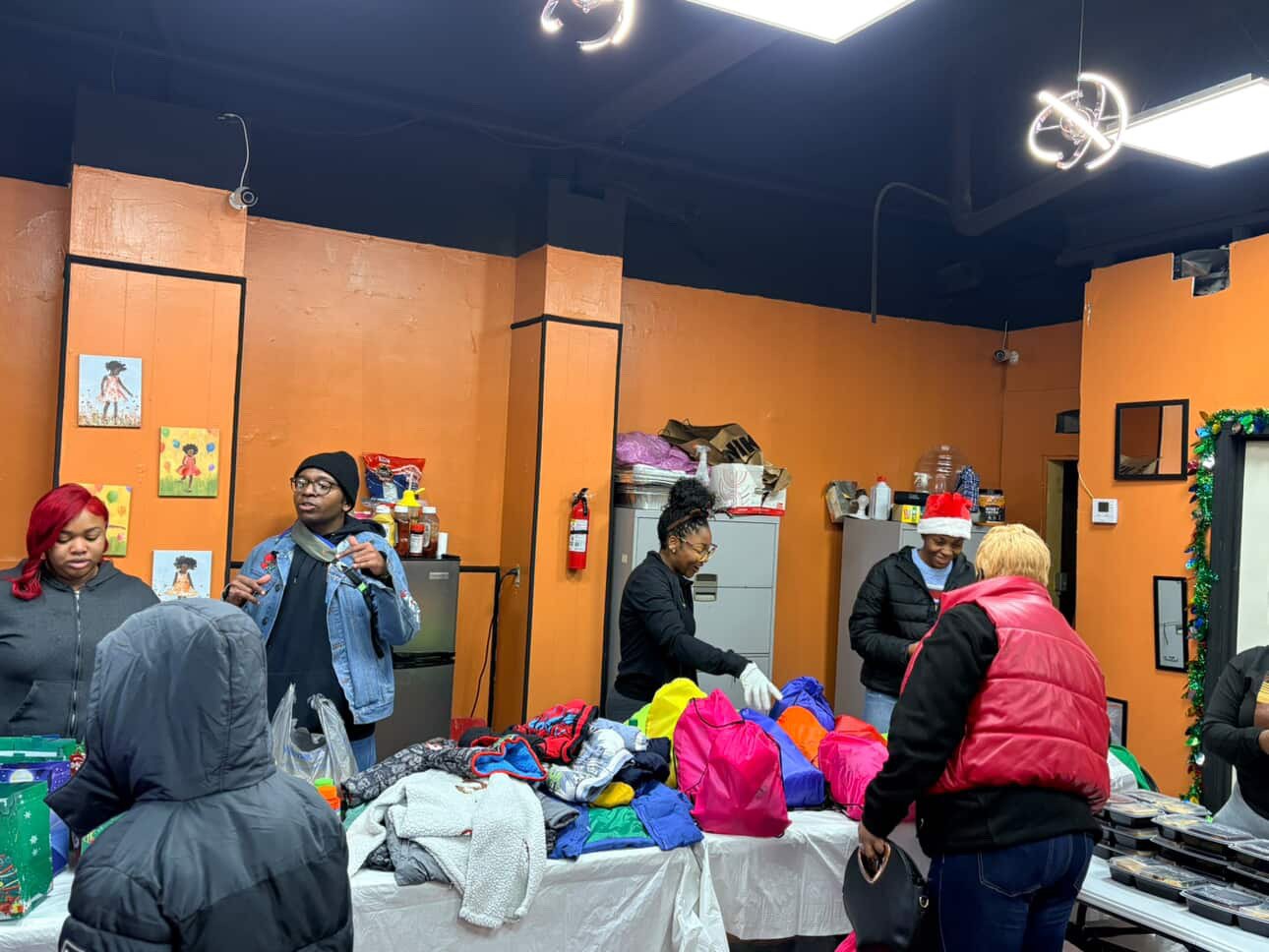 People shopping at a holiday gift sale with tables of colorful clothing and accessories, some wearing winter jackets and one person wearing a Santa hat.