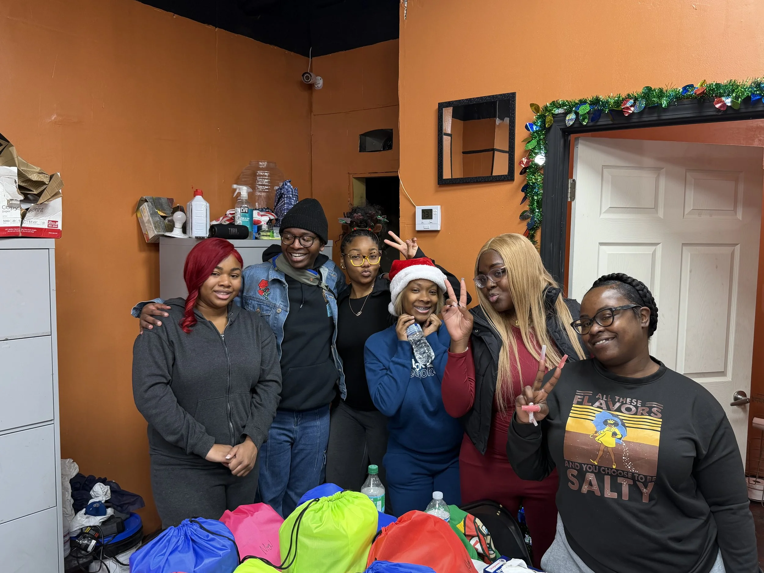 A group of six women posing together indoors, some making peace signs, with a decorated Christmas door in the background.
