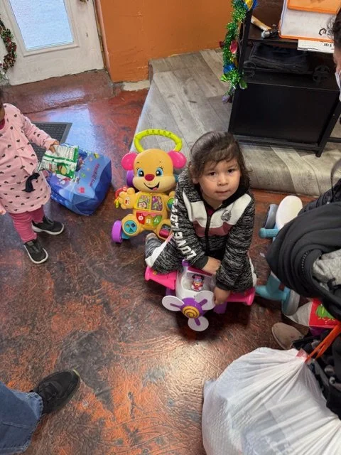 A young girl sitting on a pink and purple toy ride-on with a bear face design, surrounded by other children and toys in a room with brown flooring and a small decorated Christmas tree.