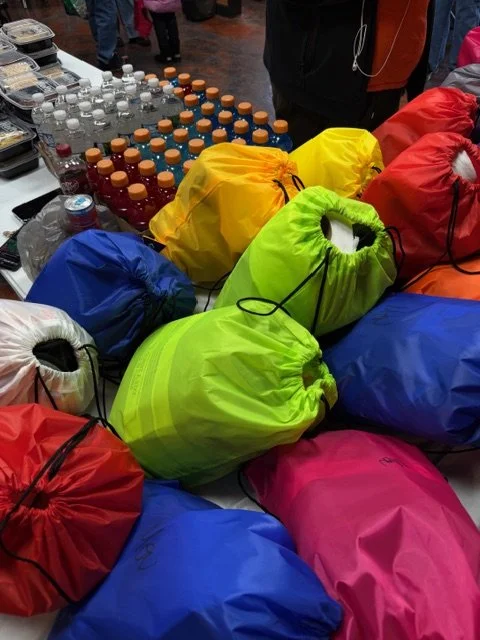 Colorful drawstring bags in yellow, green, red, blue, and pink arranged on a table alongside bottles of flavored drinks or water.
