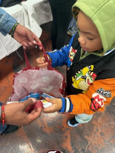 A young boy receiving a small item, possibly a snack or toy, from an adult's hands. The boy is wearing a colorful Super Mario jacket and a lime green hoodie, with a focused expression as he looks at the item. An adult is holding a clear plastic bag w